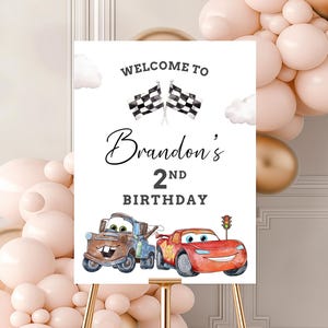 May include: A white sign on a gold easel with the text "Welcome to Brandon's 2nd Birthday." The sign features illustrations of race cars and checkered flags, with a backdrop of pink and gold balloons.