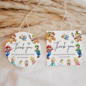 Puede incluir: Dos etiquetas de regalo blancas con temática de Mario. Una es redonda, la otra cuadrada. Ambas presentan personajes de Mario y el texto "Thank you". Las etiquetas también incluyen el mensaje "We hope you had a Mario Time! Love, Brandon."