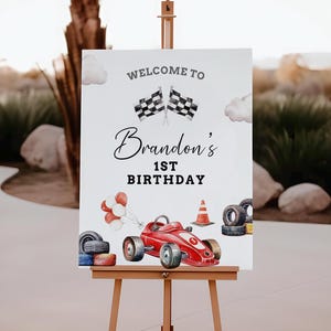 May include: Welcome to Brandon's 1st Birthday sign featuring a red racing car, chequered flags, and balloons. The sign is on an easel.
