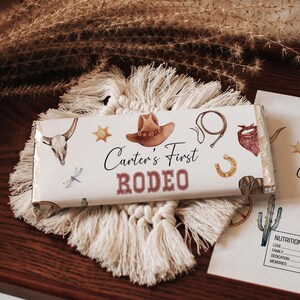 May include: A chocolate bar wrapper with a rodeo theme, featuring illustrations of a cowboy hat, horseshoe, and a steer skull. The text "Carter's First Rodeo" is prominently displayed. The wrapper rests on a macrame coaster.