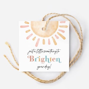 Puede incluir: Una etiqueta de regalo blanca con un diseño de sol acuarela en tonos de rosa, naranja y amarillo. La etiqueta tiene el texto "just a little something to brighten your day!" en letras de colores.