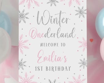 May include: A white sign with pink and silver snowflakes and glitter text. The sign says "Winter Wonderland" and "Welcome to Emilia's 1st Birthday".