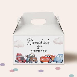 Może przedstawiać: Białe pudełko z uchwytem, ozdobione napisem "Brandon's 1st Birthday" i ilustracjami samochodów z kreskówek. Pudełko jest ozdobione chmurami. Samochody to czerwona ciężarówka, brązowy holownik, czerwony samochód wyścigowy i niebieski samochód.