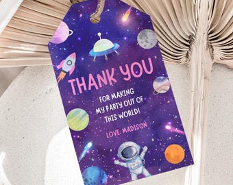 May include: A purple gift tag with a space theme. The tag features illustrations of planets, a rocket ship, and an astronaut. The text "THANK YOU" is in pink, with the message "FOR MAKING MY PARTY OUT OF THIS WORLD! LOVE: MADISON."