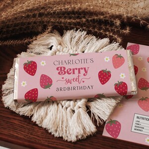 May include: A pink candy bar wrapper with the text "Charlotte's Berry Sweet 3rd Birthday" and strawberry illustrations. The candy bar is on a white macrame coaster, with a pink card featuring strawberries.