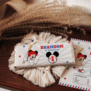 May include: A chocolate bar wrapper and a card with a baseball-themed design featuring Mickey Mouse. The text "BRANDON IS TURNING" is displayed in red and blue. The design includes stars and a baseball.