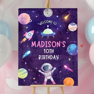 May include: A purple and black space-themed welcome sign with a cartoon astronaut, planets, stars, and a rocket ship. The sign says "Welcome to Madison's 10th Birthday".