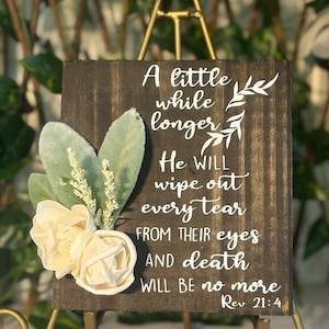 May include: A wooden sign with the text "A little while longer..." and a floral arrangement. The sign is dark brown with white lettering and features cream-colored roses and green leaves. The text includes "Rev 21:4".