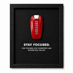 Puede incluir: Una pieza de arte mural enmarcada que presenta una llave de coche Ferrari roja centrada sobre un fondo negro. Debajo de la llave, el texto dice: STAY FOCUSED. STAY FOCUSED, STAY CONSISTENT, AND SUCCESS WILL FOLLOW. El marco es negro con acabado texturizado.