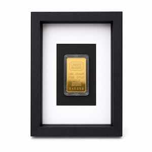 Può includere: Un lingotto d'oro fino Credit Suisse da un'oncia con purezza 999,9 e numero di serie 840599, racchiuso in una custodia protettiva in plastica trasparente ed esposto all'interno di una cornice in legno nero con bordo passe-partout bianco.