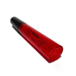 May include: A red lip gloss tube with a black cap. The tube has a label that says "7 Smiles" and "By Ari" with smiley faces.