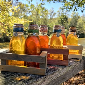 May include: Six glass jars filled with golden honey, each sealed with a cork and colorful twine. The jars are displayed in wooden crates, with a natural outdoor backdrop. Each jar has a sunflower charm.