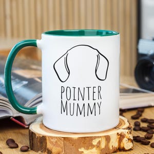 以下が含まれることがあります： グリーンの持ち手と内側を備えた白いセラミックマグ。犬の耳の黒いアウトラインと「POINTER MUMMY」の文字が特徴です。マグカップは木製のコースターの上に置かれ、コーヒー豆が散らばっています。