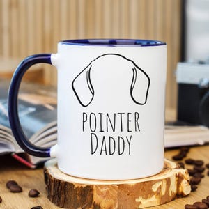 May include: White ceramic mug with a blue interior and handle, featuring a black outline of a dog's head and the text "POINTER DADDY". The mug sits on a wooden coaster, surrounded by coffee beans.
