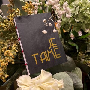 Peut inclure: Une carte noire avec l'inscription dorée "JE T'AIME" est insérée dans un bouquet de fleurs. Les fleurs comprennent des fleurs blanches, jaunes et vertes. La carte est reliée avec du fil rouge.