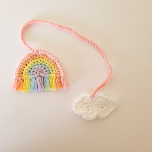 Op de afbeelding: Een regenboogkleurig gehaakt bladwijzer met een witte gehaakte wolk eraan bevestigd. De bladwijzer is gemaakt van garen en heeft een roze touwtje.