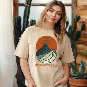 Könnte beinhalten: Ein beiges T-Shirt mit einem Grafikdruck einer Bergkette in Grün- und Blautönen. Die Grafik ist von einem roten Kreis mit dem Text "God Needs Nothing" umgeben, und der Text "He Wants You" befindet sich unterhalb der Grafik.