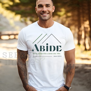 May include: White t-shirt with the word "ABIDE" in dark teal, a house graphic, and the text "You'll never find a home like Him. John 15:9 II" in green and black. The shirt is worn by a person outdoors.
