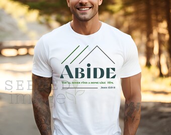 以下が含まれることがあります： 「ABIDE」の文字がダークティール色で、家のグラフィックと「You'll never find a home like Him. John 15:9 II」のテキストが緑と黒でプリントされた白いTシャツ。屋外で人が着ています。