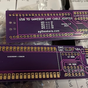 May include: Two purple circuit boards with gold-colored connectors. The top board has the text "USB TO GAMEBOY LINK CABLE ADAPTER" and the text "agtbaskara.com". The bottom board has the text "USB PORT" and the text "6182989AP-230620".