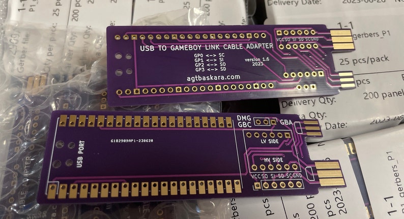 May include: Two purple circuit boards with gold-colored connectors. The top board has the text "USB TO GAMEBOY LINK CABLE ADAPTER" and the text "agtbaskara.com". The bottom board has the text "USB PORT" and the text "6182989AP-230620".