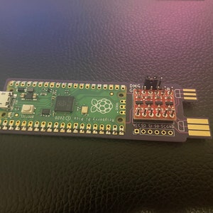 May include: A Raspberry Pi Pico microcontroller board with a green circuit board and a purple edge. The board has a USB connector, a button, and a Raspberry Pi logo. A small red circuit board with a header connector is attached to the microcontroller board. The text "Raspberry Pi Pico 2020" and "BOOTSEL" are printed on the board.