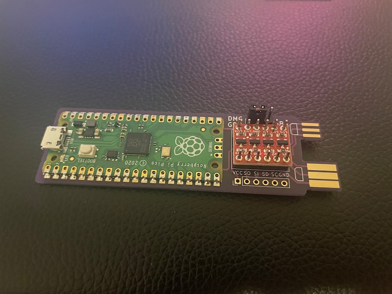 May include: A Raspberry Pi Pico microcontroller board with a green circuit board and a purple edge. The board has a USB connector, a button, and a Raspberry Pi logo. A small red circuit board with a header connector is attached to the microcontroller board. The text "Raspberry Pi Pico 2020" and "BOOTSEL" are printed on the board.