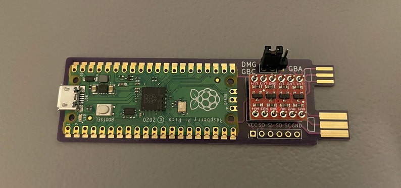 May include: A green Raspberry Pi Pico board with a USB-C port and gold-colored pins. A red board with gold-colored pins is connected to the Raspberry Pi Pico board. The red board has the text "DMG GBC GBA" and "VCC SO SI SD-SCGND" printed on it.