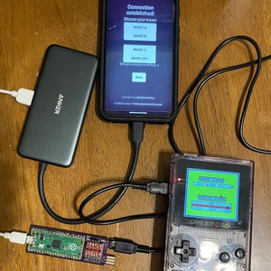 May include: A close-up of a Game Boy Color handheld gaming console connected to a Raspberry Pi computer. The Game Boy Color is transparent and has a blue screen displaying the text "GAME TYPE" and "MUSIC TYPE". The Raspberry Pi is connected to a power bank and a smartphone.