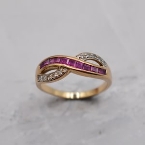 May include: A gold-toned ring with an infinity symbol design. The ring features a row of square-cut, vibrant pink gemstones and small, clear gemstones. The band has a polished finish, and the gemstones are set in a channel setting.