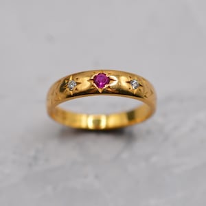 Op de afbeelding: Een gouden ring met een centrale rode edelsteen en twee kleinere witte edelstenen. De ring heeft een band met geëtste ster- en bloemmotieven. De ring wordt getoond op een lichtgrijze ondergrond.