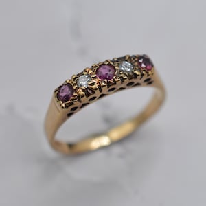 May include: A gold ring with five gemstones. Alternating round rubies and diamonds are set in the ring's band. The rubies are a deep pink colour, and the diamonds are clear. The ring has an openwork design along the band. The background is a soft, neutral colour.