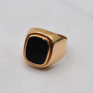 May include: A gold-toned signet ring featuring a smooth, polished finish and a rectangular black inlay centered on the face. The band is solid and tapers slightly toward the back, showcasing a clean, metallic surface with a reflective quality.