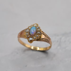 Può includere: Un anello in oro con un opale ovale iridescente al centro, circondato da un gruppo di piccole perle bianche. La fascia presenta un motivo inciso e testurizzato lungo i lati, che prosegue in un gambo in oro liscio e lucido.