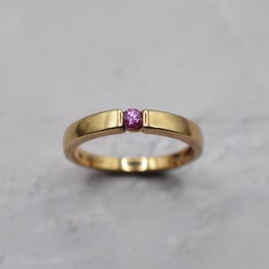 Op de afbeelding: Een gouden bandring met een enkele, kleine, rond geslepen roze edelsteen in een spanningzetting. Het metaal heeft een gepolijste, reflecterende afwerking en de ring behoudt een constante breedte rond de vinger, wat de centrale steen benadrukt.