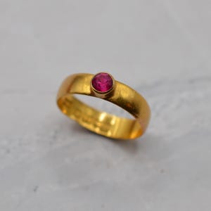 May include: A gold band ring featuring a single, round, vibrant pink gemstone set in a bezel setting. The metal surface of the ring has a matte, brushed finish, and the interior of the band shows visible stamped markings on the inner surface.