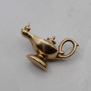 Może przedstawiać: Mała zawieszka z metalu w kolorze złotym w kształcie lampy oliwnej. Zawieszka posiada zaokrąglony korpus, wąski dzióbek z małą pionową końcówką, okrągłą podstawę oraz zakrzywiony uchwyt z boku. Powierzchnia ma gładkie, polerowane wykończenie.