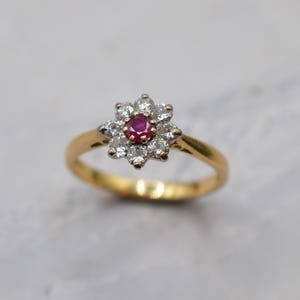Könnte beinhalten: Ein Goldring mit einem zentralen, rund geschliffenen roten Rubin, der von acht kleinen, klaren, rund geschliffenen weißen Diamanten in einer blumenartigen Anordnung umgeben ist. Die Fassung der Steine ist silberfarben und bildet einen Kontrast zum Goldring.