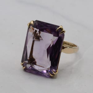 Può includere: Un anello in oro con una grande pietra preziosa ametista viola, rettangolare e sfaccettata, tenuta in posizione da quattro griffe in oro. La fascia è realizzata in oro giallo lucido e si collega alla montatura alla base della pietra preziosa.