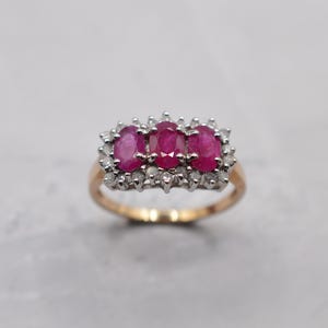 May include: A gold ring featuring three oval-cut rubies, each surrounded by a halo of small, clear gemstones. The ring's band is a warm gold color, and the gemstones are a vibrant red.