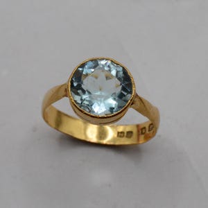 May include: A gold ring featuring a round, faceted, light blue gemstone set in a bezel setting. The band is smooth and polished gold, with visible hallmarks stamped on the inner surface of the ring band, indicating its metal purity and origin.