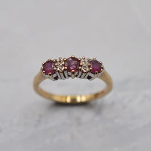 Peut inclure: Une bague en or ornée de trois pierres précieuses rouges de taille ovale disposées en rangée, alternées avec deux petites pierres claires. Le sertissage des pierres est argenté, contrastant avec l'anneau en or poli et lisse.