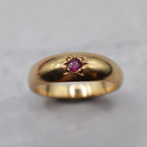 May include: A gold band ring featuring a smooth, rounded surface with a single, small, round-cut pink gemstone set in the center. The gemstone is surrounded by an engraved starburst pattern that radiates outward from the stone into the gold metal.