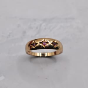 Puede incluir: Un anillo dorado con tres gemas magenta en forma de estrella. El anillo tiene una banda lisa y redondeada. Las gemas están espaciadas uniformemente en la parte superior del anillo.
