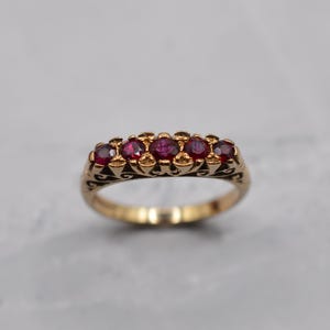 May include: A gold ring with five round, deep red gemstones. The ring has an ornate, filigree design along the band, with decorative swirls and patterns. The gemstones are set closely together, creating a continuous line of colour.