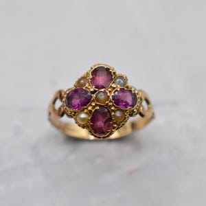 May include: A gold ring featuring a cluster of four oval-cut deep pink gemstones arranged in a cross pattern, interspersed with four small round white pearls. The metal band has a decorative openwork design on the sides near the central setting.