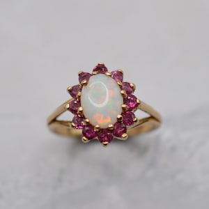 May include: A gold ring featuring a central oval-shaped white opal surrounded by a halo of twelve small, round, deep pink gemstones. The band is split at the shoulders and connects to the central setting, which holds the opal and pink stones.