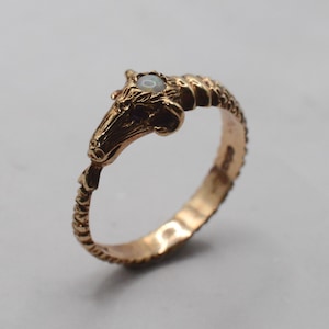 May include: A gold ring featuring a sculpted ram head design with a small, round, iridescent opal stone set between the horns. The band of the ring has a textured, ribbed pattern that extends from the ram head around the entire circumference.