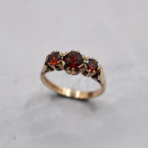 Op de afbeelding: Een goudkleurige ring met drie ronde, dieprode edelstenen. De ring heeft een sierlijk bandontwerp en de edelstenen zijn gezet in een klassieke, elegante stijl. De ring wordt gepresenteerd op een lichtgrijze ondergrond.