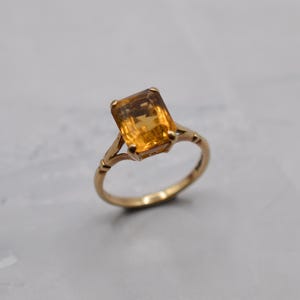 May include: A gold ring featuring a rectangular, amber-colored gemstone. The ring has a delicate, ornate design with a slender band. The gemstone is the focal point, catching the light and showcasing its warm, golden hue. A classic piece of jewelry.